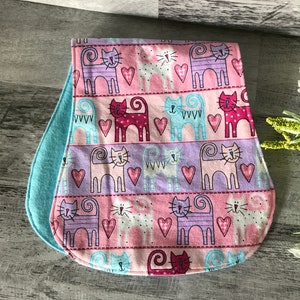 May include: A pink and blue fabric burp cloth with a pattern of cats and hearts. The fabric is soft and absorbent.