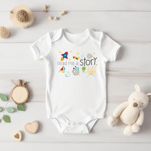 May include: White baby onesie with short sleeves and a colorful graphic that says "read me a story". The graphic includes a rocket, a bird, a ship, a whale, a tree, and a castle.