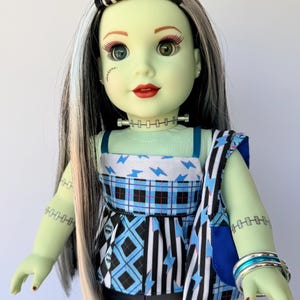 May include: A doll with light green skin, long black hair with white streaks, and a stitched scar on the cheek. The doll wears a sleeveless top with blue lightning bolts and plaid patterns, a matching shoulder bag, and black pants with silver bracelets.
