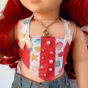 May include: A doll wearing a red and white seashell-themed top with a gold necklace and denim shorts. The top has a heart-shaped design with pearl accents and small bows. The doll has red hair and a flower hair accessory.