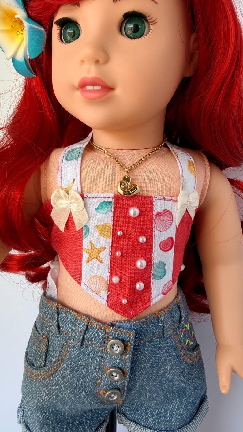 May include: A doll wearing a red and white seashell-themed top with a gold necklace and denim shorts. The top has a heart-shaped design with pearl accents and small bows. The doll has red hair and a flower hair accessory.