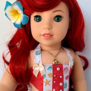 May include: A doll with bright red hair and green eyes, wearing a red and white seashell-themed top and denim shorts. A blue and yellow flower adorns the hair. A gold necklace with a seashell pendant is also visible.