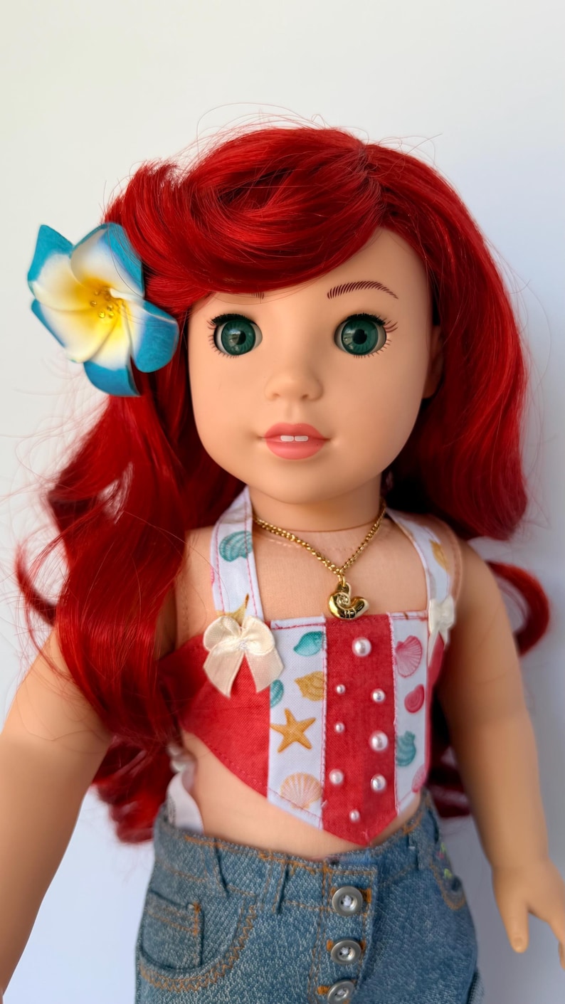 May include: A doll with bright red hair and green eyes, wearing a red and white seashell-themed top and denim shorts. A blue and yellow flower adorns the hair. A gold necklace with a seashell pendant is also visible.