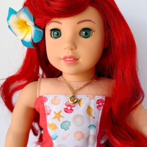 May include: A doll with red hair and green eyes, wearing a dress with a seashell pattern. A blue and yellow flower adorns the doll's hair. A gold necklace with a seashell pendant is visible. The doll is a children's toy.