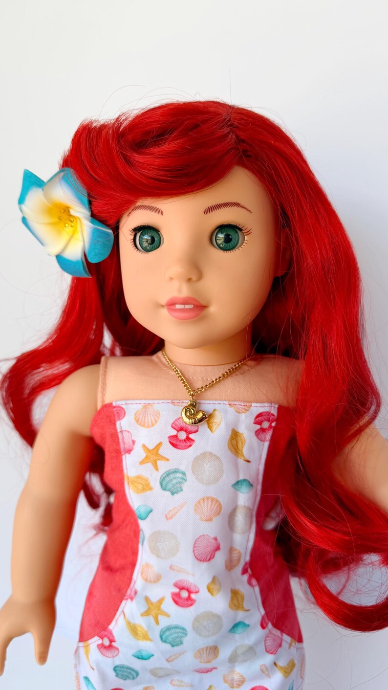 May include: A doll with red hair and green eyes, wearing a dress with a seashell pattern. A blue and yellow flower adorns the doll's hair. A gold necklace with a seashell pendant is visible. The doll is a children's toy.