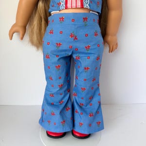 Könnte beinhalten: Ein zweiteiliges Puppenoutfit. Das Oberteil ist weiß mit roten und blauen Akzenten. Die Hose ist hellblau mit einem roten Blumenmuster. Die Puppe trägt rote Schuhe.