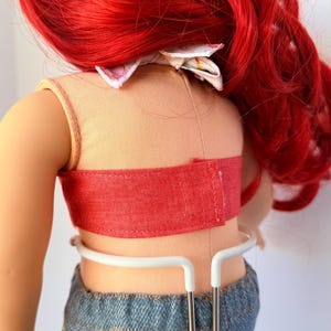 May include: A doll with bright red hair, wearing a peach-colored top with a red band and denim shorts. The doll is held by a white metal stand. A floral bow is at the back of the doll's head.