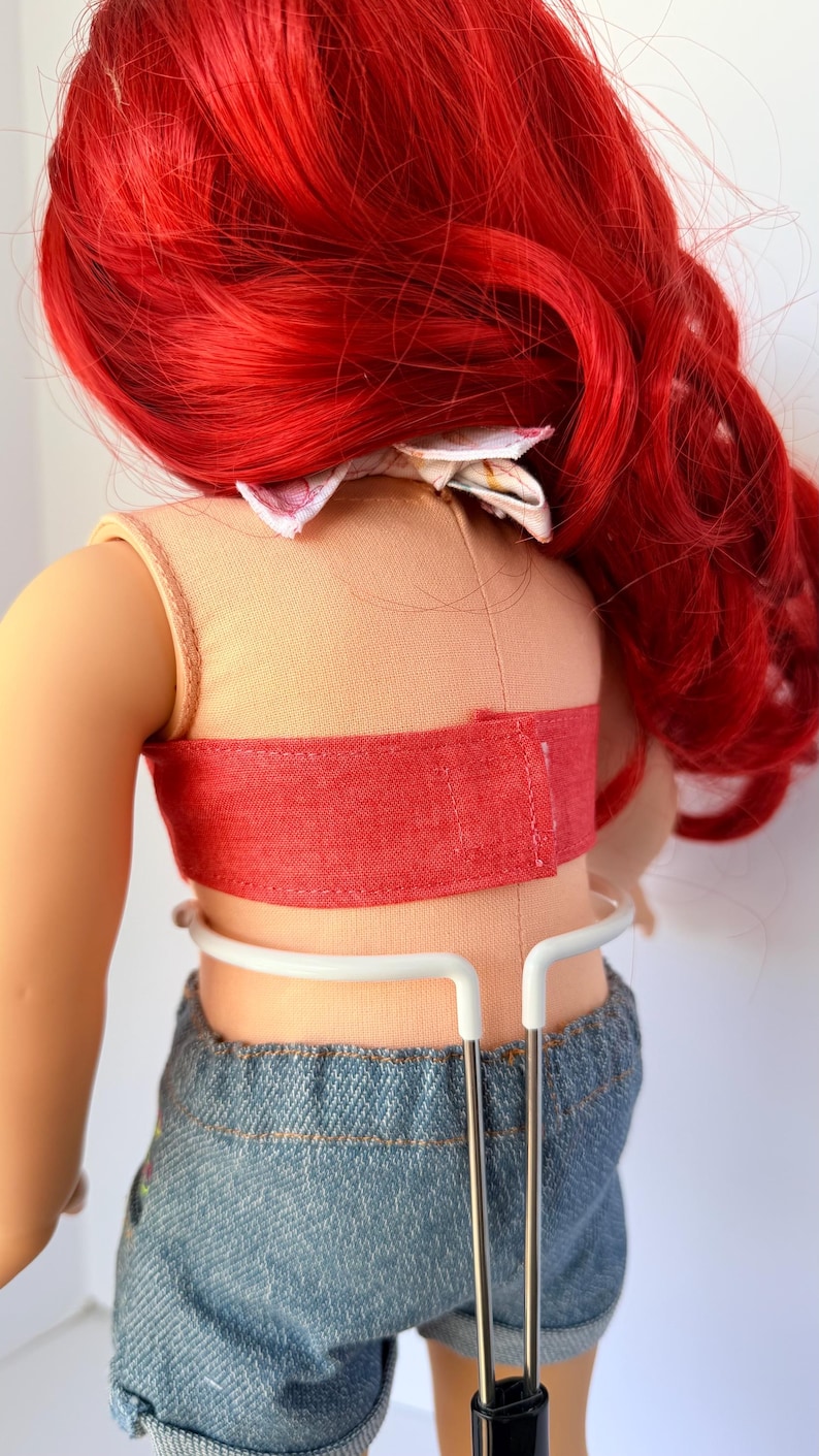 May include: A doll with bright red hair, wearing a peach-colored top with a red band and denim shorts. The doll is held by a white metal stand. A floral bow is at the back of the doll's head.