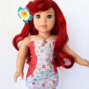 May include: A doll with red hair and green eyes, wearing a seashell-patterned dress with a red accent and a white lace hem. A blue and yellow flower adorns the doll's hair. A gold necklace is also visible.