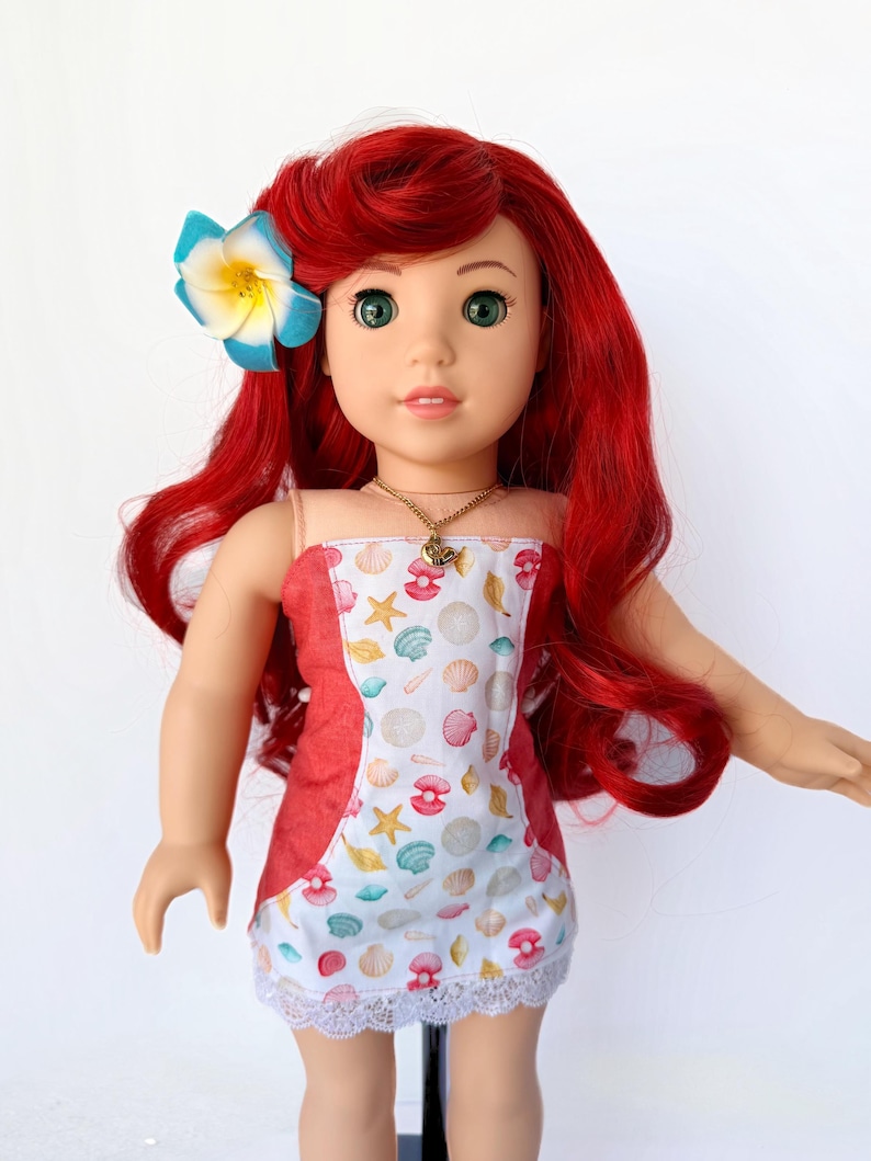May include: A doll with red hair and green eyes, wearing a seashell-patterned dress with a red accent and a white lace hem. A blue and yellow flower adorns the doll's hair. A gold necklace is also visible.