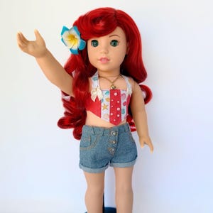 May include: A doll with red hair, green eyes, and a blue and yellow flower in her hair. She wears a red and white patterned top, denim shorts, a gold necklace, and pink sandals. The doll is standing on a blue stand.