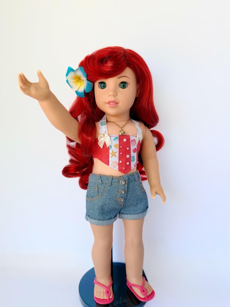 May include: A doll with red hair, green eyes, and a blue and yellow flower in her hair. She wears a red and white patterned top, denim shorts, a gold necklace, and pink sandals. The doll is standing on a blue stand.