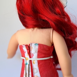 May include: A doll wearing a red strapless dress with a seashell pattern and a white lace trim. The doll has bright red hair and a white bow. The dress has a zipper down the back.