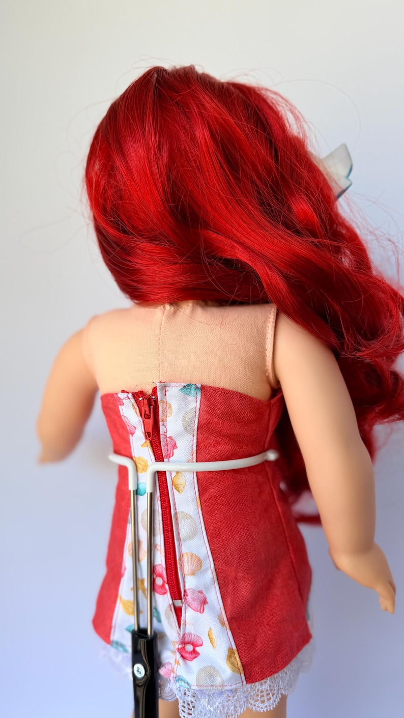 May include: A doll wearing a red strapless dress with a seashell pattern and a white lace trim. The doll has bright red hair and a white bow. The dress has a zipper down the back.