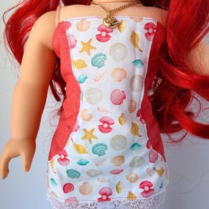 May include: A doll wearing a red and white sundress with a seashell pattern and a white lace hem. The dress has a red trim and thin straps. A gold necklace with a seashell pendant is around the doll's neck. The doll has red hair.