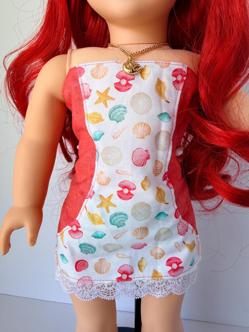 May include: A doll wearing a red and white sundress with a seashell pattern and a white lace hem. The dress has a red trim and thin straps. A gold necklace with a seashell pendant is around the doll's neck. The doll has red hair.