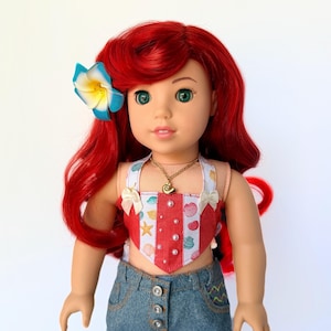 May include: A doll with red hair, green eyes, and a blue and yellow flower hair accessory. The doll wears a red and white seashell-themed top, a gold necklace, and denim jeans. The doll is a children's toy.