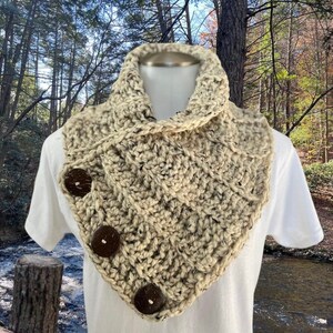 May include: A beige crocheted cowl with a textured pattern and three brown wooden buttons. The cowl is worn on a white t-shirt.