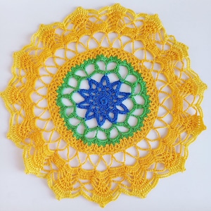 May include: A handmade, circular doily with a vibrant color scheme. The doily features a central blue starburst design, surrounded by a green ring, and a large yellow outer layer. The crochet work is intricate and detailed, showcasing a delicate, openwork pattern.