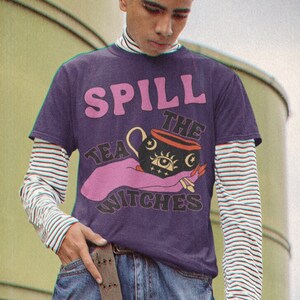 May include: A purple t-shirt with a graphic of a pink hand holding a black cauldron with a single eye and the text "Spill the Tea Witches".