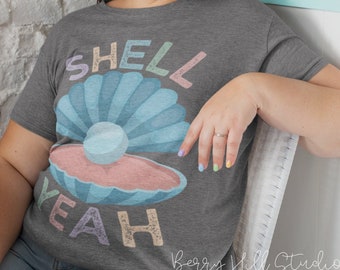 Può includere: Una maglietta grigia con una grafica colorata di una conchiglia con una perla all'interno. Il testo "SHELL YEAH" è scritto in un font arcobaleno.