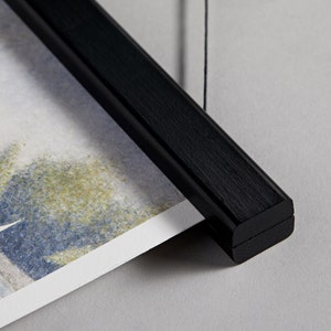 May include: Black wooden hanging frame with a string attached to the top. The frame is holding a watercolour print of a landscape.