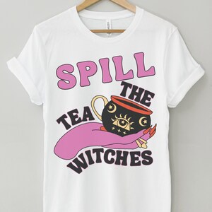 May include: White t-shirt with a pink and black graphic design that says "Spill the Tea Witches". The graphic features a cauldron with a crescent moon, stars, and an eye.