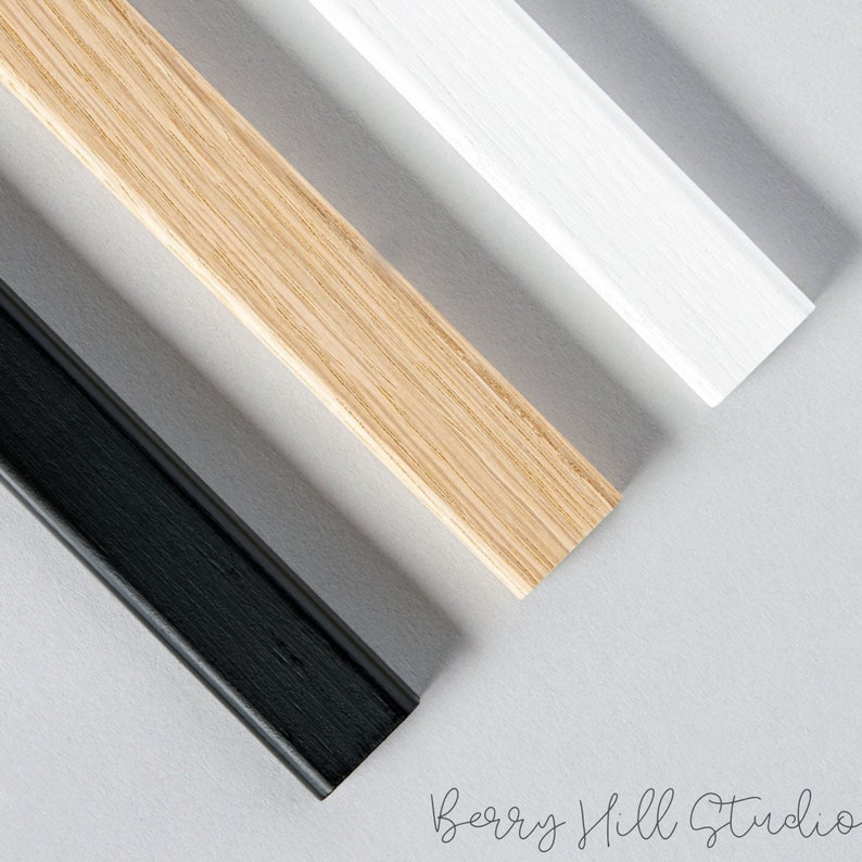 May include: Three wooden picture frames in different colors: natural wood, white, and black. The frames are arranged in a diagonal line on a gray background.