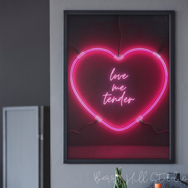 May include: A pink neon heart-shaped sign with the text "love me tender" inside. The sign is framed in black and hanging on a gray wall.