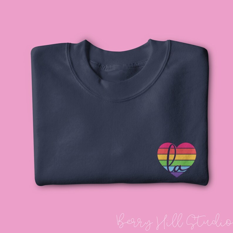 Pu&ograve; includere: Una felpa blu navy con un design a forma di cuore arcobaleno e la lettera "L" al suo interno. Il cuore &egrave; composto da strisce di rosso, arancione, giallo, verde, blu, indaco e viola.