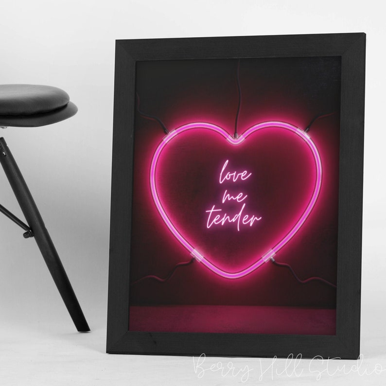 May include: A black framed print with a pink neon heart that says "love me tender" on a dark red background.