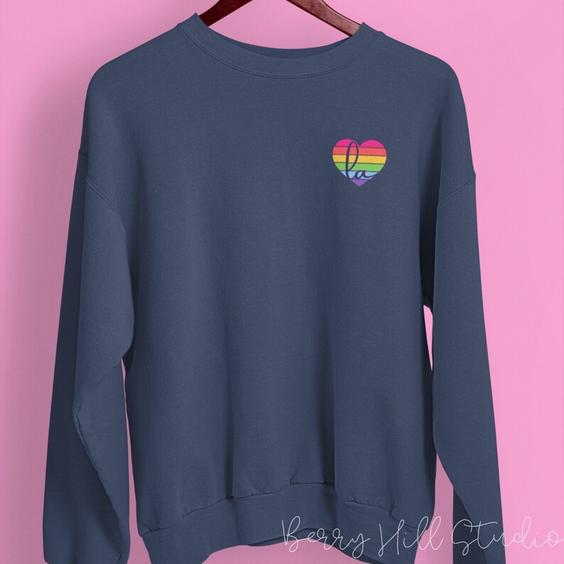 Pu&ograve; includere: Una felpa blu navy con un cuore arcobaleno sul petto sinistro. Il cuore contiene le lettere "la" all'interno.