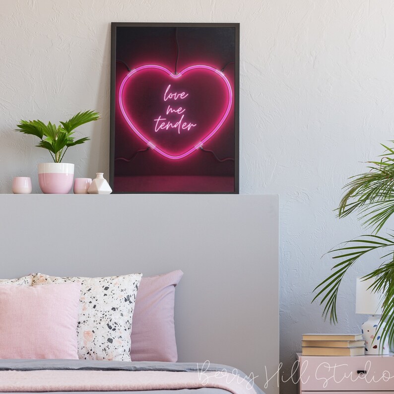 May include: A framed print of a pink neon heart sign with the text "love me tender" on a black background. The print is hanging on a gray wall above a bed with pink and white pillows.
