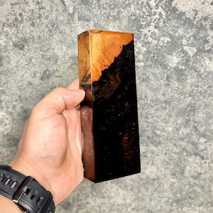 Puede incluir: Un bloque rectangular de madera con un llamativo contraste de colores. La parte superior es de un marrón claro cálido, que pasa a un marrón oscuro profundo. La veta de la madera es visible, añadiendo textura. El bloque se sostiene en una mano.