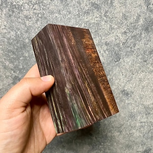 Könnte beinhalten: Ein Holzblock mit einem dunkelbraunen, violetten und grünen Farbmuster. Das Holz hat eine natürliche Maserung und ist glatt.