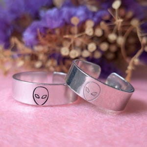 Puede incluir: Dos anillos ajustables plateados con un diseño de cara alienígena grabada. Los anillos son de metal brillante. El fondo es rosa suave con flores moradas borrosas, creando un contraste.