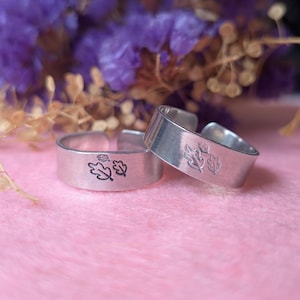 May include: Two silver adjustable rings with leaf designs. The rings are made of brushed metal and set against a soft pink background with purple flowers. The rings have a simple, elegant design, suitable for everyday wear.