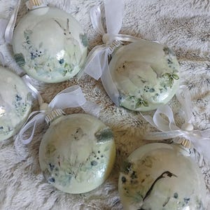 May include: Several round Christmas ornaments with white ribbon hangers. Each ornament features a different animal illustration, including a rabbit and a bird, surrounded by floral and botanical elements in shades of green and blue.