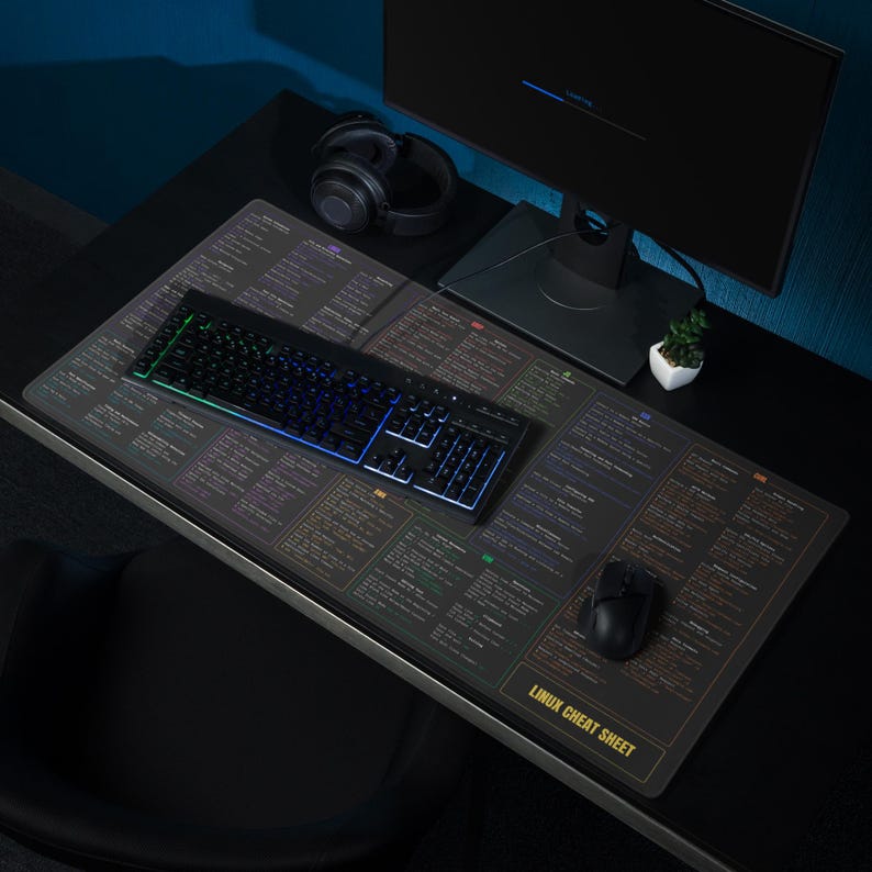 May include: A black computer keyboard with multi-colored LED lights sits on a large mouse pad. The mouse pad has a dark background with a printed Linux cheat sheet. A computer monitor, headphones, and mouse are also on the desk.