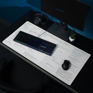 May include: A white desk mat with a grid pattern and text printed on it. A black keyboard with rainbow backlighting, a black mouse, and a pair of black headphones are on the mat. A potted plant and a computer monitor are on the desk.