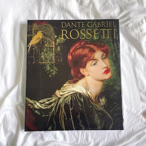 May include: A hardcover book titled "Dante Gabriel Rossetti" featuring a portrait of a woman with red hair and a gold-trimmed black gown. A yellow bird sits in a cage on the cover, with the title in gold lettering.