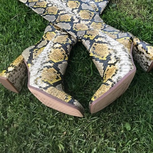 May include: A pair of knee-high boots with a snakeskin pattern in shades of yellow, gray, and black. The boots have a block heel and are resting on green grass.