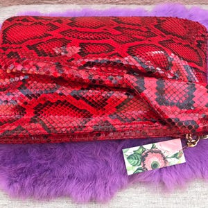 May include: A red clutch with a snakeskin pattern, featuring black and red scales. The clutch is rectangular with a flap closure and a gold chain. It rests on a purple, fluffy surface.