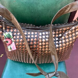 Op de afbeelding: Een geweven leren handtas in brons- en donkerbruine tinten. Het heeft lange, olijfgroene leren riemen en een decoratief label met bloemmotieven. De tas staat op een turquoise ondergrond.