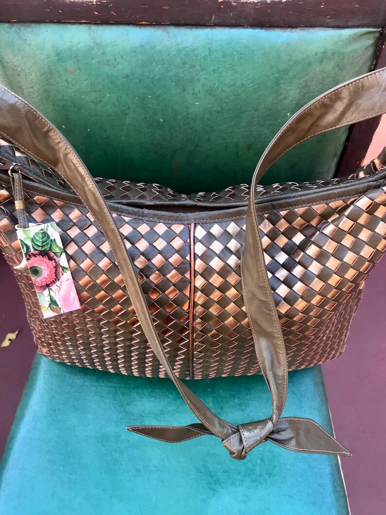 Op de afbeelding: Een geweven leren handtas in brons- en donkerbruine tinten. Het heeft lange, olijfgroene leren riemen en een decoratief label met bloemmotieven. De tas staat op een turquoise ondergrond.