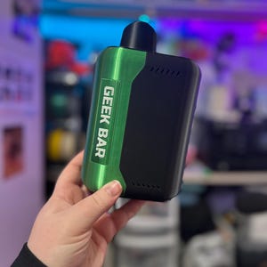 Peut inclure: Un appareil de vapotage Geek Bar noir et vert. L'appareil a un panneau vert avec le texte "GEEK BAR" en blanc. L'appareil a un dessus noir et un corps noir. L'appareil est tenu dans une main.