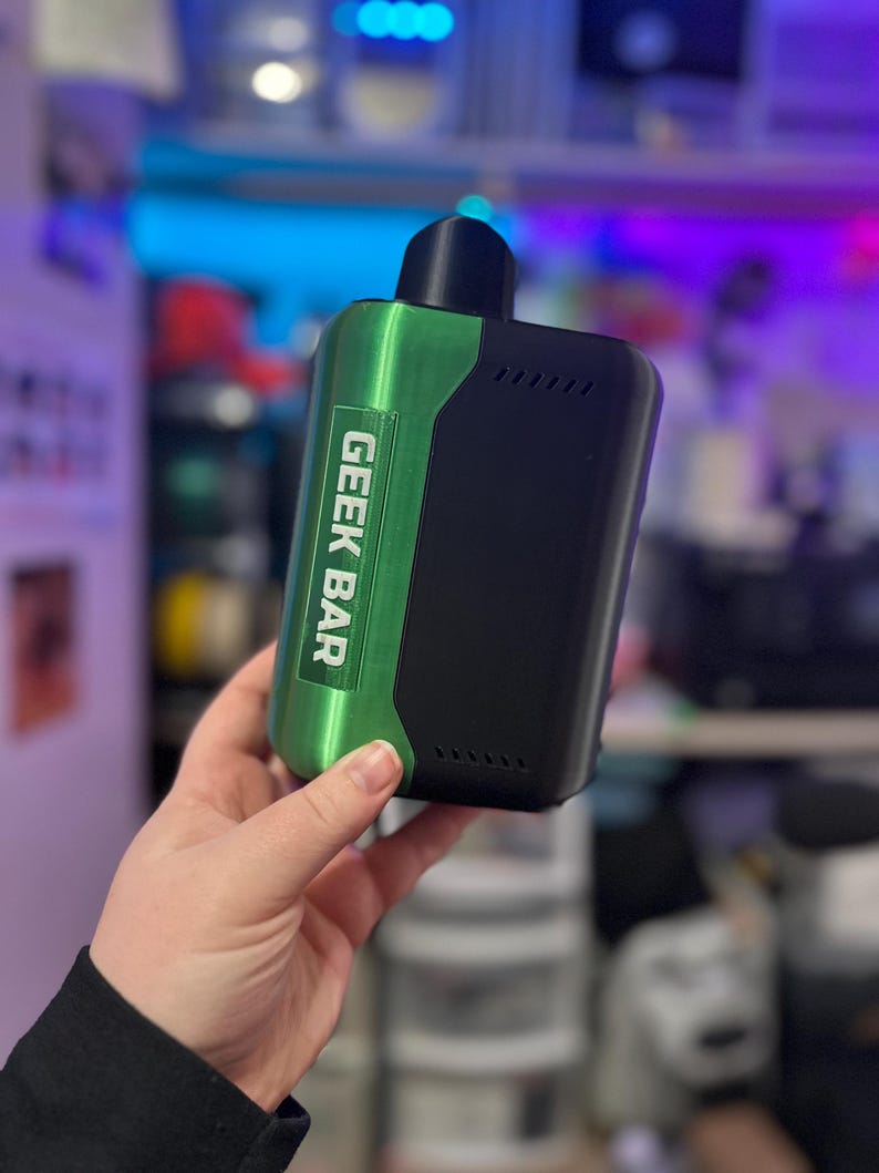 Peut inclure: Un appareil de vapotage Geek Bar noir et vert. L'appareil a un panneau vert avec le texte "GEEK BAR" en blanc. L'appareil a un dessus noir et un corps noir. L'appareil est tenu dans une main.