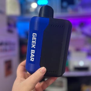 Peut inclure: Un appareil de vapotage Geek Bar bleu et noir. L'appareil porte les mots "GEEK BAR" imprim&eacute;s verticalement en blanc sur une section bleue. L'appareil est tenu dans une main et poss&egrave;de un embout buccal noir.