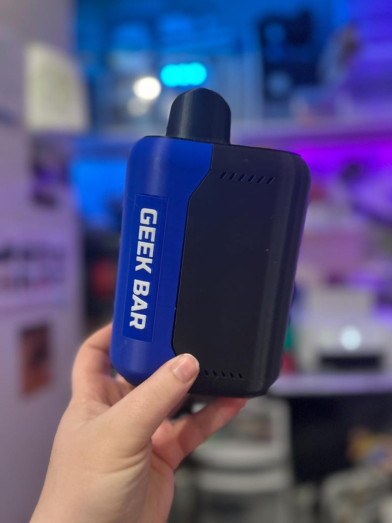 Peut inclure: Un appareil de vapotage Geek Bar bleu et noir. L'appareil porte les mots "GEEK BAR" imprim&eacute;s verticalement en blanc sur une section bleue. L'appareil est tenu dans une main et poss&egrave;de un embout buccal noir.