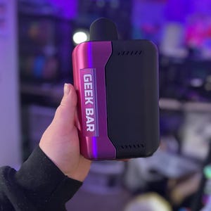Peut inclure: Un appareil de vapotage Geek Bar. L'appareil a un corps noir avec un accent m&eacute;tallique violet. Le texte "GEEK BAR" est imprim&eacute; verticalement sur le c&ocirc;t&eacute; violet. L'appareil a un embout buccal noir.